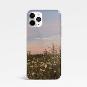 May include: Transparent phone case displaying a sunset scene with a field of white flowers. The text "to live for the hope of it all" is printed on the case. The case is designed to fit a smartphone.