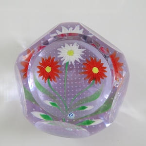 May include: A clear glass paperweight with a faceted exterior and a lavender-tinted interior. The center features a floral arrangement with two red flowers, one white flower, green stems, and leaves, all surrounded by tiny air bubbles.