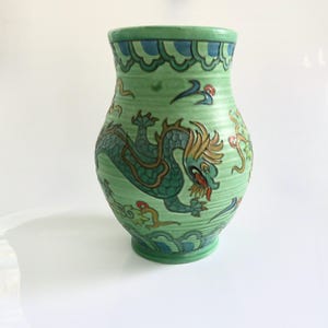 May include: A green ceramic vase with a dragon design. The vase features a detailed dragon illustration in shades of green, blue, and gold. The vase has a wide mouth and a textured surface, with decorative patterns around the rim.