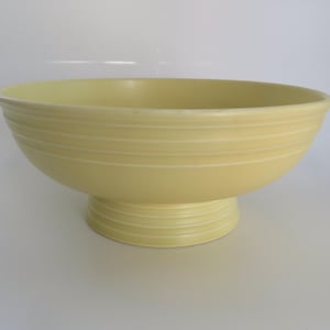 May include: A pale yellow ceramic bowl with a pedestal base. The bowl has a wide, rounded shape with horizontal ridged detailing around the exterior. The pedestal base also features similar ridged detailing. The bowl is likely for serving or display.