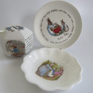 May include: A collection of white ceramic items featuring illustrations from Beatrix Potter's stories. Includes a plate with a rim, a scalloped dish, and a multi-sided piggy bank. The plate has text around the rim.