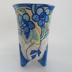 May include: A ceramic vase with a three-legged base, featuring a floral design in shades of blue, green, and tan. The vase has a textured, off-white body and a flared top. The blue flowers are the focal point of the design.