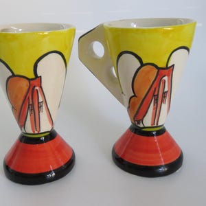May include: Two ceramic mugs with a unique design. The mugs feature a yellow rim, white body with orange and red accents, and a red base with a black trim. Each mug has a handle with a circular opening.