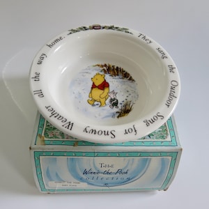 May include: A white ceramic bowl featuring a central illustration of Winnie the Pooh and Piglet in the snow. The rim is inscribed with the text, They sang the Outdoor Song for Snowy Weather all the way home. The bowl rests on a decorative box.
