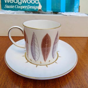 May include: A white teacup and saucer set with a gold rim and leaf design. The cup features a handle and is decorated with brown and purple leaf patterns. The saucer has a matching design and gold accents. The set is from the "Susie Cooper Design" collection.