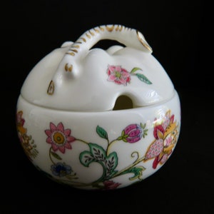 May include: A white ceramic sugar bowl with a lid and handle, decorated with vibrant floral patterns. The bowl features pink, yellow, and blue flowers with green leaves. The handle and rim have gold detailing. The bowl is round and set against a black backdrop.