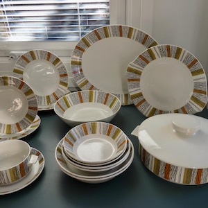 Op de afbeelding: Een verzameling wit keramisch serviesgoed met een modern design uit het midden van de eeuw. De borden, kommen en serveerschaal hebben een gestreepte rand in bruin, groen en grijs. Inclusief borden, kommen en een serveerschaal.
