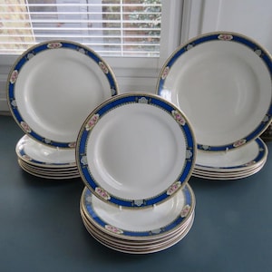 Pode incluir: Um conjunto de pratos de cerâmica branca com uma borda decorativa azul e dourada. A borda apresenta um design floral com detalhes em rosa e branco. Os pratos são empilhados, mostrando um conjunto de jantar clássico.
