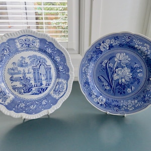 May include: Two blue and white decorative plates. One plate features a scenic landscape design, the other a floral pattern. Both have scalloped edges and are displayed on stands. The plates are in the style of antique china.