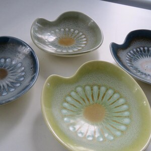 May include: Four ceramic dishes in shades of blue, green, and yellow. Each dish has a unique shape with a scalloped edge and a central floral design in a contrasting color. The dishes are arranged on a white surface.