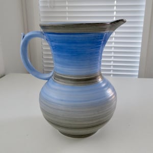 May include: A ceramic pitcher with a blue and gray striped design. The pitcher has a handle and a spout. The colors transition from light blue to gray. The pitcher is a decorative item.