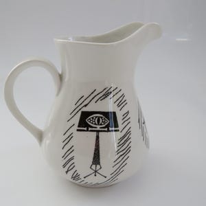 May include: A white ceramic pitcher featuring a black graphic illustration of a music stand with an eye motif, surrounded by abstract hatched lines. The pitcher has a curved handle and a pouring spout, with a smooth, glossy finish throughout.