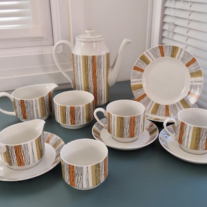 May include: A vintage tea set featuring a white teapot, cups, saucers, plates, and creamers. The set has a mid-century modern design with vertical stripes in orange, brown, and gold. The set is on a blue surface.