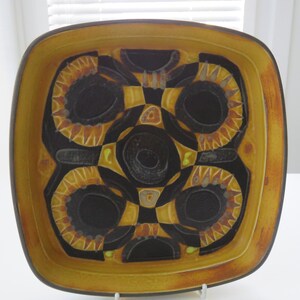 Peut inclure: Plat carré en céramique avec un motif géométrique. Le plat présente un centre noir avec des accents jaunes et orange, et un bord jaune. Le motif comprend des formes circulaires et carrées.