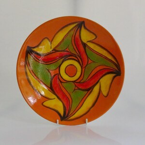 Può includere: Un piatto rotondo in ceramica con uno sfondo arancione lucido caratterizzato da un cerchio giallo centrale circondato da un motivo astratto e vorticoso di forme rosse, verdi e gialle delineate in marrone scuro, creando un effetto visivo dinamico.
