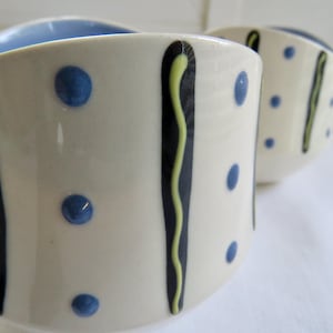 May include: Two ceramic bowls with a cream-colored exterior, blue interiors, and blue polka dots. Each bowl has a black vertical stripe with a yellow, wavy line. The bowls are likely for serving food or as decorative items.