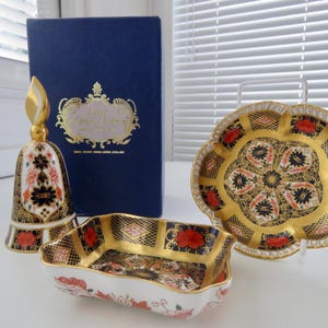 Może przedstawiać: Zestaw dekoracyjnych przedmiotów porcelanowych, w tym dzwonek, talerz i mała prostokątna miska. Elementy posiadają złote wykończenia, kwiatowe wzory i granatowe tło. Obecna jest również granatowa książka.
