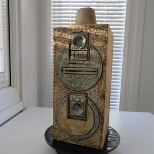 May include: A rectangular, textured ceramic vase with a cylindrical top and a circular base. The vase features two large, circular, metallic-looking designs with geometric details. The vase is tan with brown and silver accents. The vase is on a black circular base.