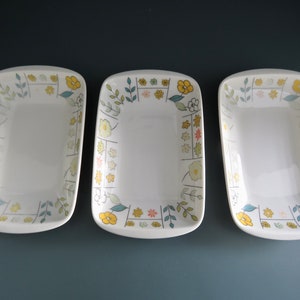 Può includere: Tre piatti da portata rettangolari in ceramica bianca con angoli arrotondati. Ogni piatto è decorato con un motivo floreale giallo, verde e rosa. I piatti sono disposti uno accanto all'altro su una superficie scura.