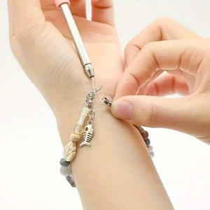 May include: A close-up of a person fastening a silver clasp on a beaded bracelet. The bracelet features a variety of beads, including white, gray, and brown, and a small silver charm shaped like a fish skeleton.
