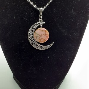 May include: A silver crescent moon pendant necklace with a pink and brown marbled stone in the centre.