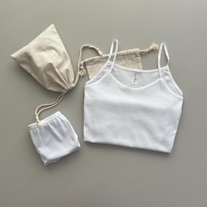 Puede incluir: Camiseta blanca de tirantes finos, cuello redondo y una pequeña etiqueta blanca en la parte delantera. La camiseta está sobre una superficie gris claro con dos bolsas de tela beige.