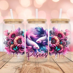 Könnte beinhalten: Drei klare Glasbecher mit Bambusdeckeln und Strohhalmen. Jeder Becher hat ein anderes Design: ein rosa und blauer Fisch mit einem Sonnenblumen- und Schmetterlingsdesign, ein rosa und blauer Fisch mit dem Wort "Pisces" und einem Sonnenblumen- und Schmetterlingsdesign sowie ein Sonnenblumen- und Schmetterlingsdesign.