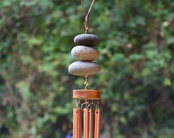 May include: A copper-colored wind chime with a stack of three gray stones at the top. The chime has several long, thin tubes that hang below a wooden disc. A square piece of copper hangs at the bottom, suspended by a chain.