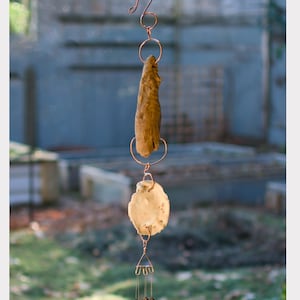 May include: A copper wind chime with a piece of driftwood, a white shell, and four brass tubes hanging from it.