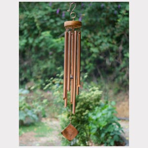 May include: A copper-colored wind chime with a wooden top and a square-shaped wooden clapper. The chime hangs from a curved metal hook. The background is a blurred view of green foliage.