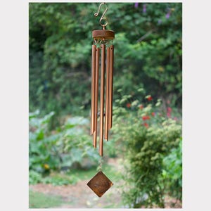 May include: A copper-colored wind chime with a wooden top and a decorative square-shaped bottom piece. The chime has several long, cylindrical tubes that hang from the wooden top. The background is a blurred view of green foliage.