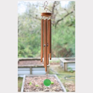 May include: A copper-colored wind chime with a wooden top and a green glass disc at the bottom. The chime has multiple cylindrical tubes of varying lengths. The background is a blurred outdoor scene with trees and garden beds.