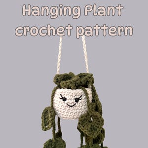 Puede incluir: Un macetero colgante de ganchillo hecho a mano con un diseño de cara sonriente. La maceta blanquecina tiene hojas verdes y una cuerda color crema para colgar. El texto "Hanging Plant crochet pattern" está en la parte superior.