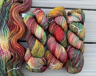 May include: Three twisted skeins of hand-dyed yarn arranged side-by-side, accompanied by a long, loose, un-twisted hank of the same fiber. The yarn features a variegated color palette of deep red, burnt orange, golden yellow, and forest green.