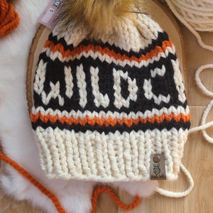 May include: White knitted hat with a black and orange striped pattern and the word "WILD" knitted in black. The hat has a fluffy brown pom pom on top and a brown leather tag with the text "Knit Wit".
