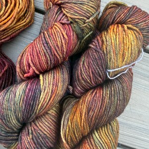 May include: Two skeins of hand-dyed yarn featuring a variegated color palette of deep burgundy, burnt orange, golden yellow, and olive green. The twisted fiber strands exhibit a multi-tonal appearance with subtle transitions between the warm autumn-inspired hues.