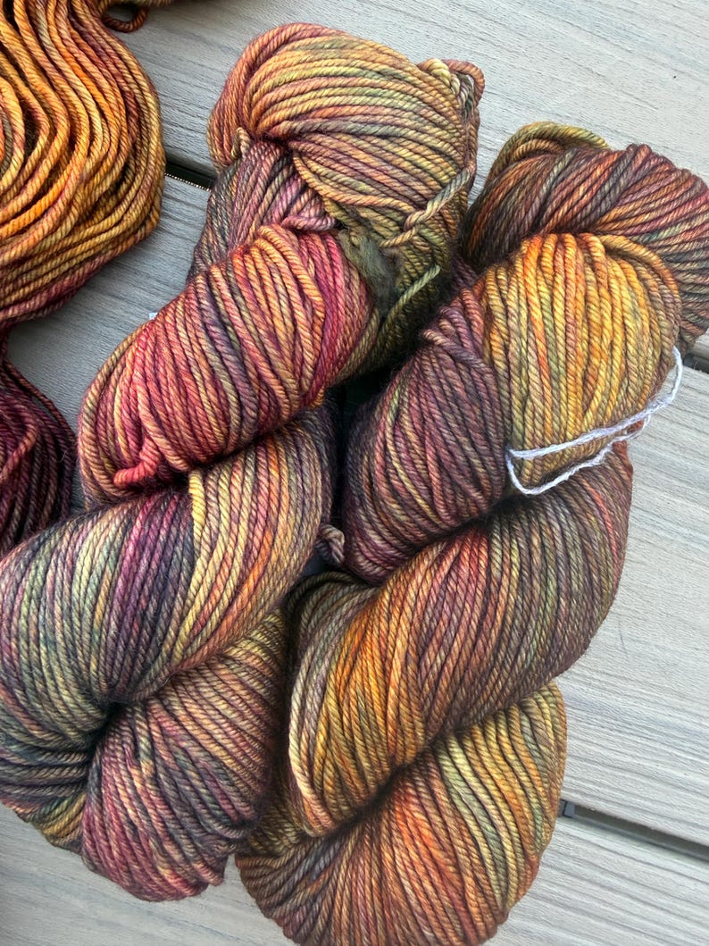 May include: Two skeins of hand-dyed yarn featuring a variegated color palette of deep burgundy, burnt orange, golden yellow, and olive green. The twisted fiber strands exhibit a multi-tonal appearance with subtle transitions between the warm autumn-inspired hues.
