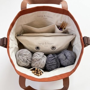 May include: An open orange tote bag with brown leather handles and accents, filled with yarn and knitting supplies. The bag has a canvas lining with pockets and a zippered pouch. The text "twig & horn" is visible on the pouch.