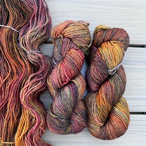 May include: Three skeins of hand-dyed yarn featuring a variegated color palette of deep forest green, burnt orange, warm mustard yellow, and dark burgundy. The yarn is twisted into two compact bundles and one long, loose, flowing hank.