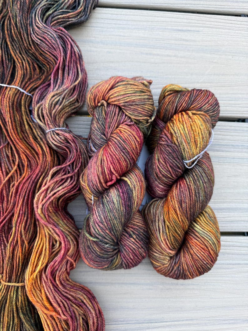 May include: Three skeins of hand-dyed yarn featuring a variegated color palette of deep forest green, burnt orange, warm mustard yellow, and dark burgundy. The yarn is twisted into two compact bundles and one long, loose, flowing hank.