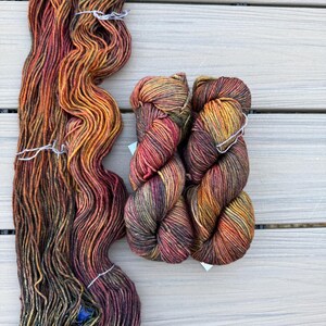 May include: One long, loose hank of yarn and two smaller, tightly twisted skeins of yarn. The yarn features a variegated color palette of deep orange, burnt red, dark brown, and olive green tones, creating a blended, multi-colored appearance.
