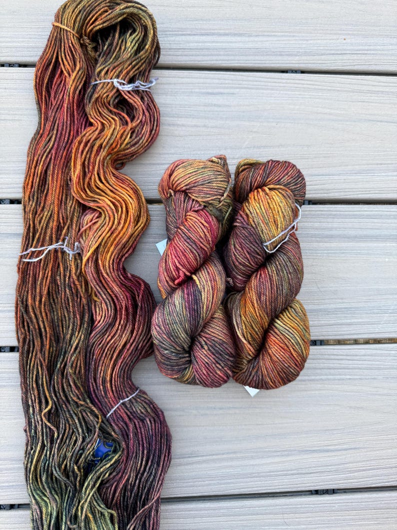 May include: One long, loose hank of yarn and two smaller, tightly twisted skeins of yarn. The yarn features a variegated color palette of deep orange, burnt red, dark brown, and olive green tones, creating a blended, multi-colored appearance.