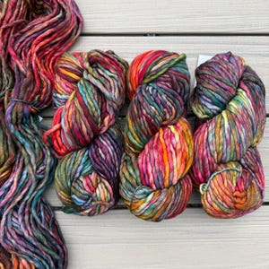 May include: A collection of thick, hand-dyed yarn skeins featuring a vibrant variegated color palette of deep magenta, bright orange, forest green, and dark purple. The yarn has a chunky, soft texture with twisted fibers throughout each skein.