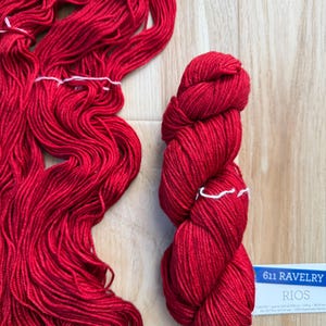 Puede incluir: Madejas y ovillos de hilo rojo brillante. Un ovillo est&aacute; cuidadosamente enrollado, mientras que el otro es suelto y fluido. Una etiqueta con el texto "611 RAVELRY RIOS" es visible.