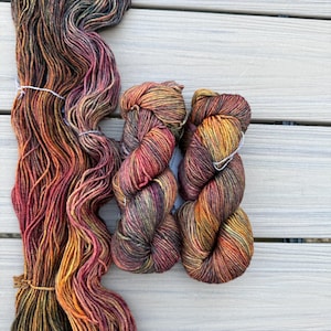 May include: Three skeins of hand-dyed yarn featuring a variegated color palette of deep forest green, burnt orange, golden yellow, and dark burgundy. The yarn is twisted into two compact bundles and one long, loose, untwisted hank of fiber.