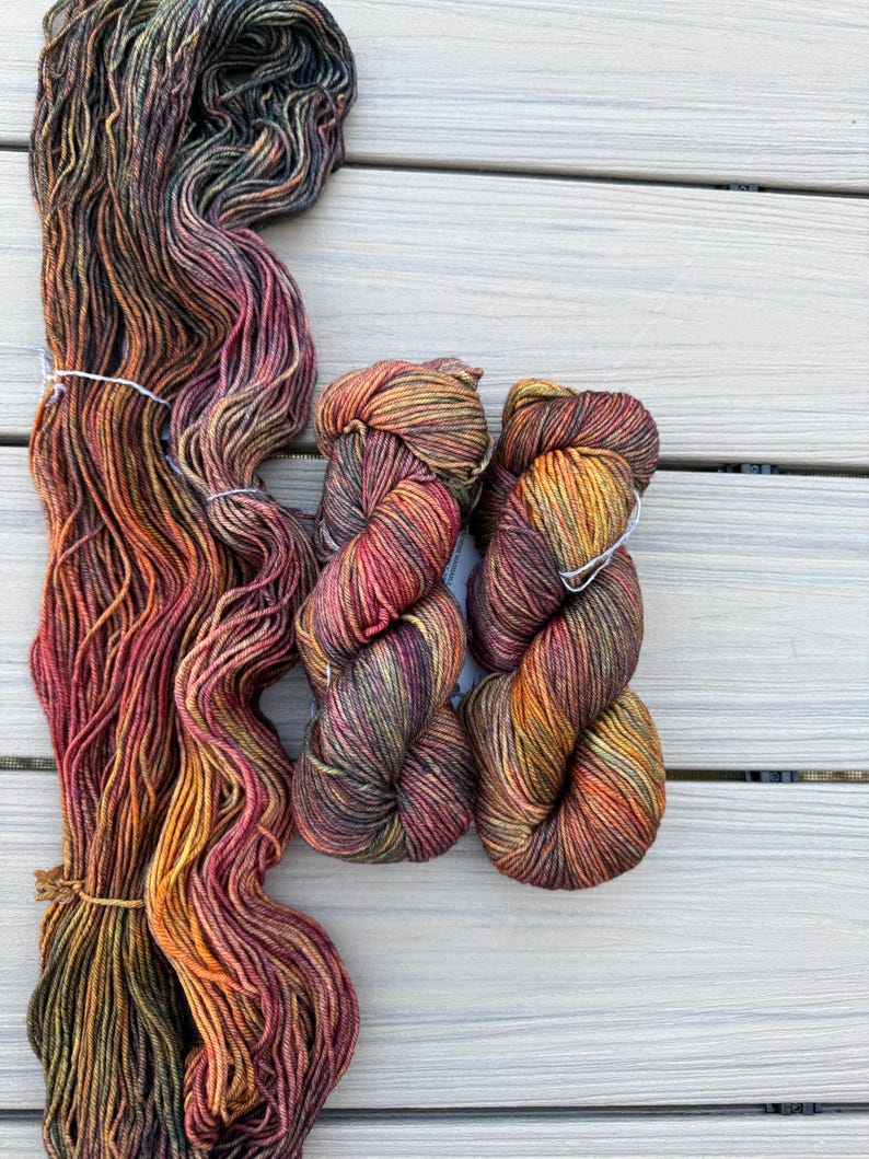 May include: Three skeins of hand-dyed yarn featuring a variegated color palette of deep forest green, burnt orange, golden yellow, and dark burgundy. The yarn is twisted into two compact bundles and one long, loose, untwisted hank of fiber.