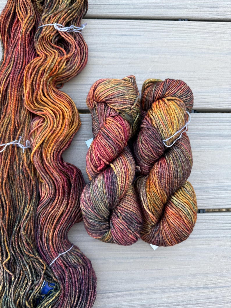 May include: Three skeins of hand-dyed yarn featuring a variegated color palette of deep burgundy, burnt orange, golden yellow, and dark forest green. One skein is loose and elongated, while the other two are tightly twisted into compact bundles.