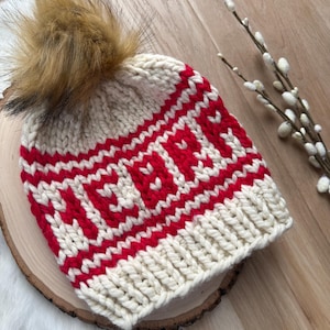 Puede incluir: Un gorro de punto color crema con letras y rayas rojas, rematado con un pompón de piel sintética marrón. El gorro se exhibe sobre una superficie de madera, con un tema invernal o festivo.