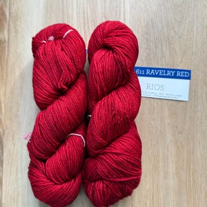 Puede incluir: Dos madejas de hilo rojo vibrante, bien enrolladas, sobre una superficie de madera clara. Una tarjeta peque&ntilde;a con el texto "611 RAVELRY RED" y "RIOS" es visible. El hilo parece suave, para tejer o hacer ganchillo.