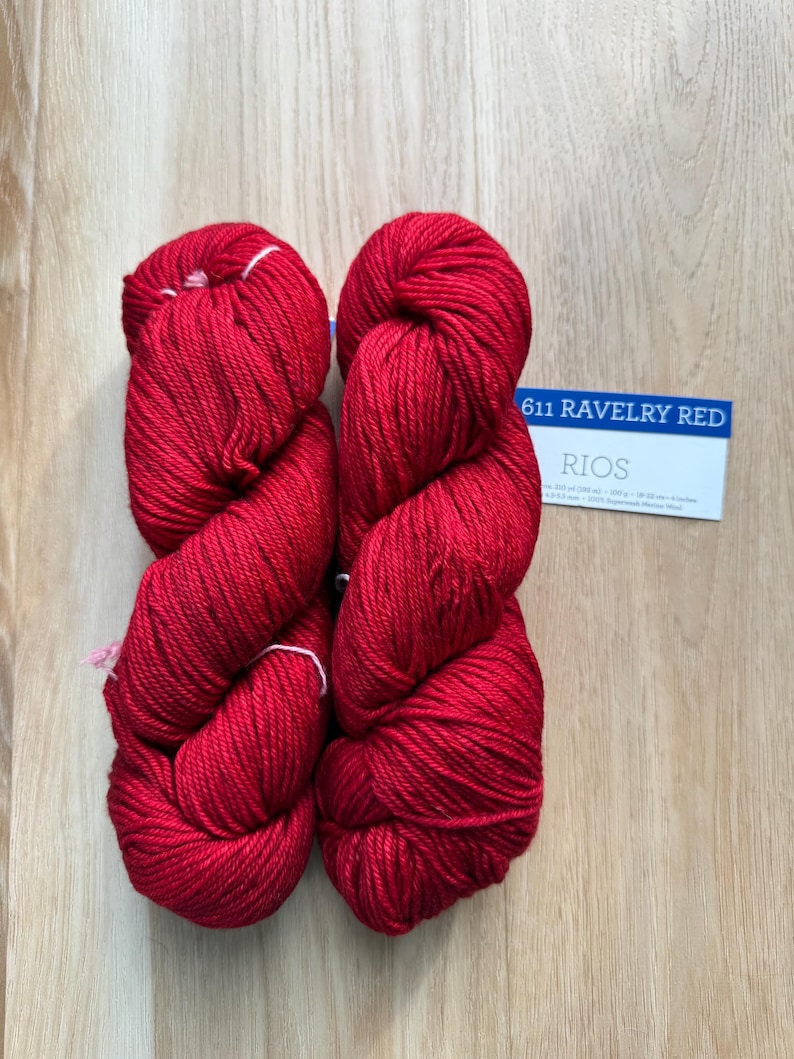 Puede incluir: Dos madejas de hilo rojo vibrante, bien enrolladas, sobre una superficie de madera clara. Una tarjeta peque&ntilde;a con el texto "611 RAVELRY RED" y "RIOS" es visible. El hilo parece suave, para tejer o hacer ganchillo.